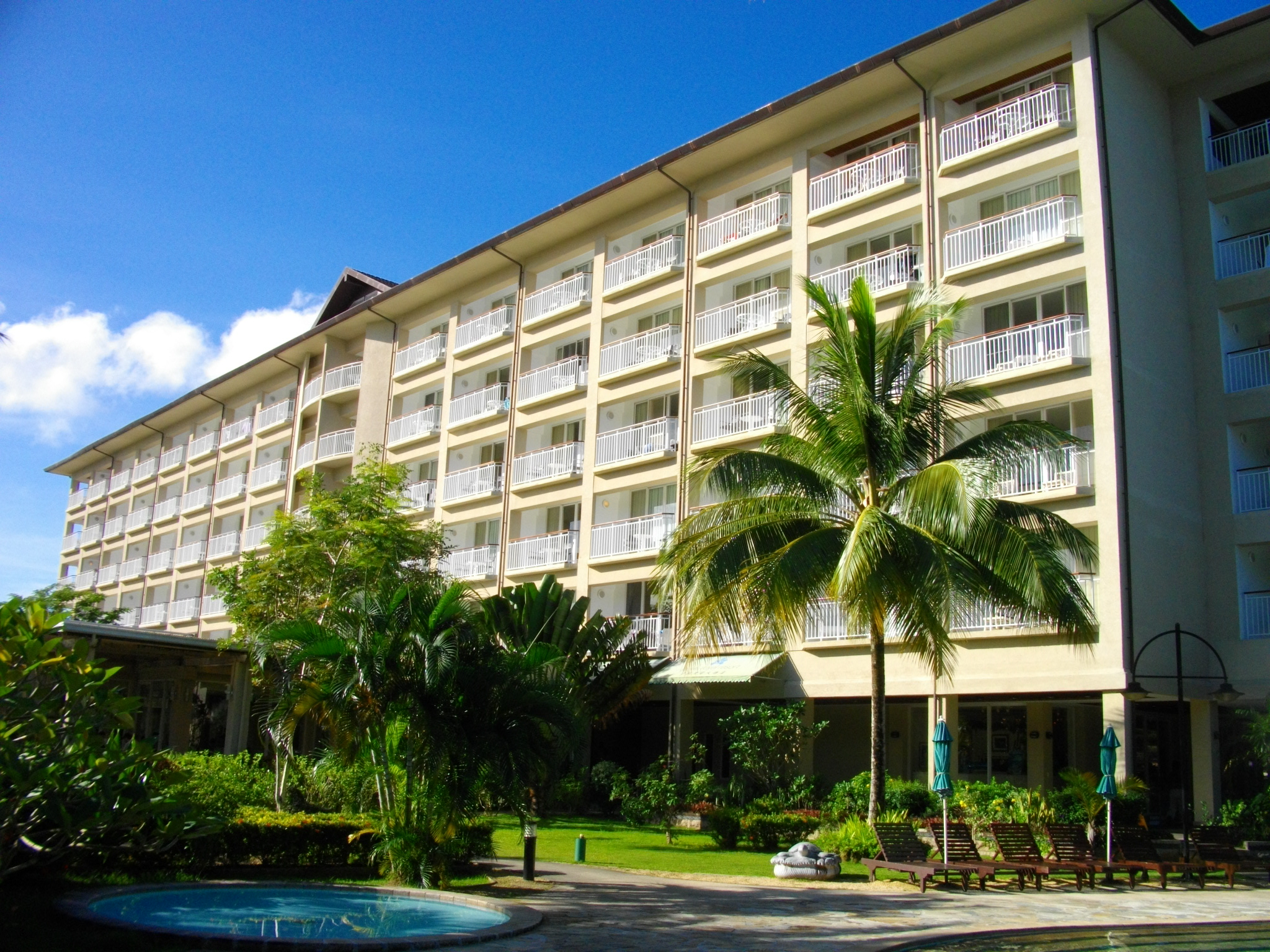 alt=Photo of a modern resort hotel building with palm trees and a swimming pool.