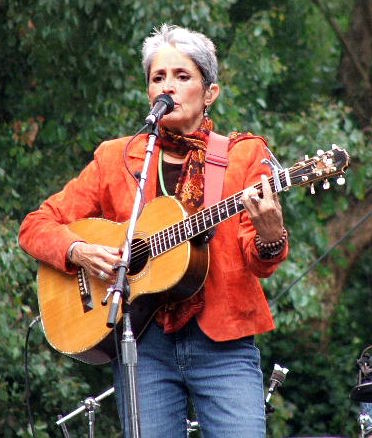 Baez plays in blue jeans and orange waist jacket, against a backdrop of lush trees