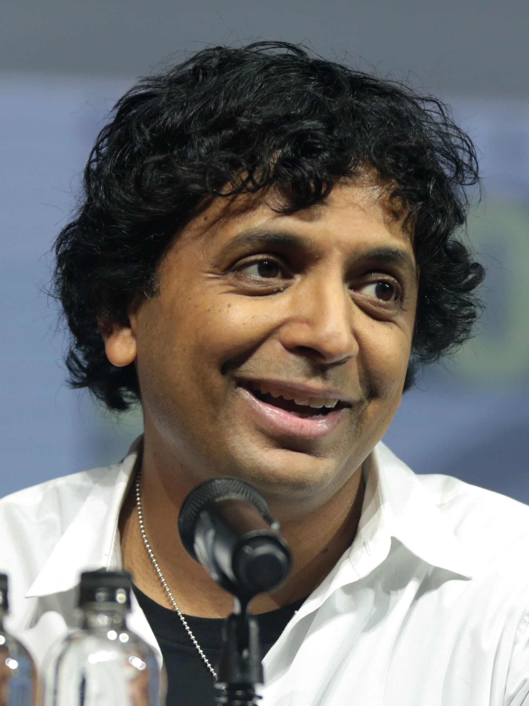 M. Night Shyamalan이 샌디에이고 코믹콘 패널에서 흰 셔츠를 입고 마이크에 대고 이야기하는 모습.