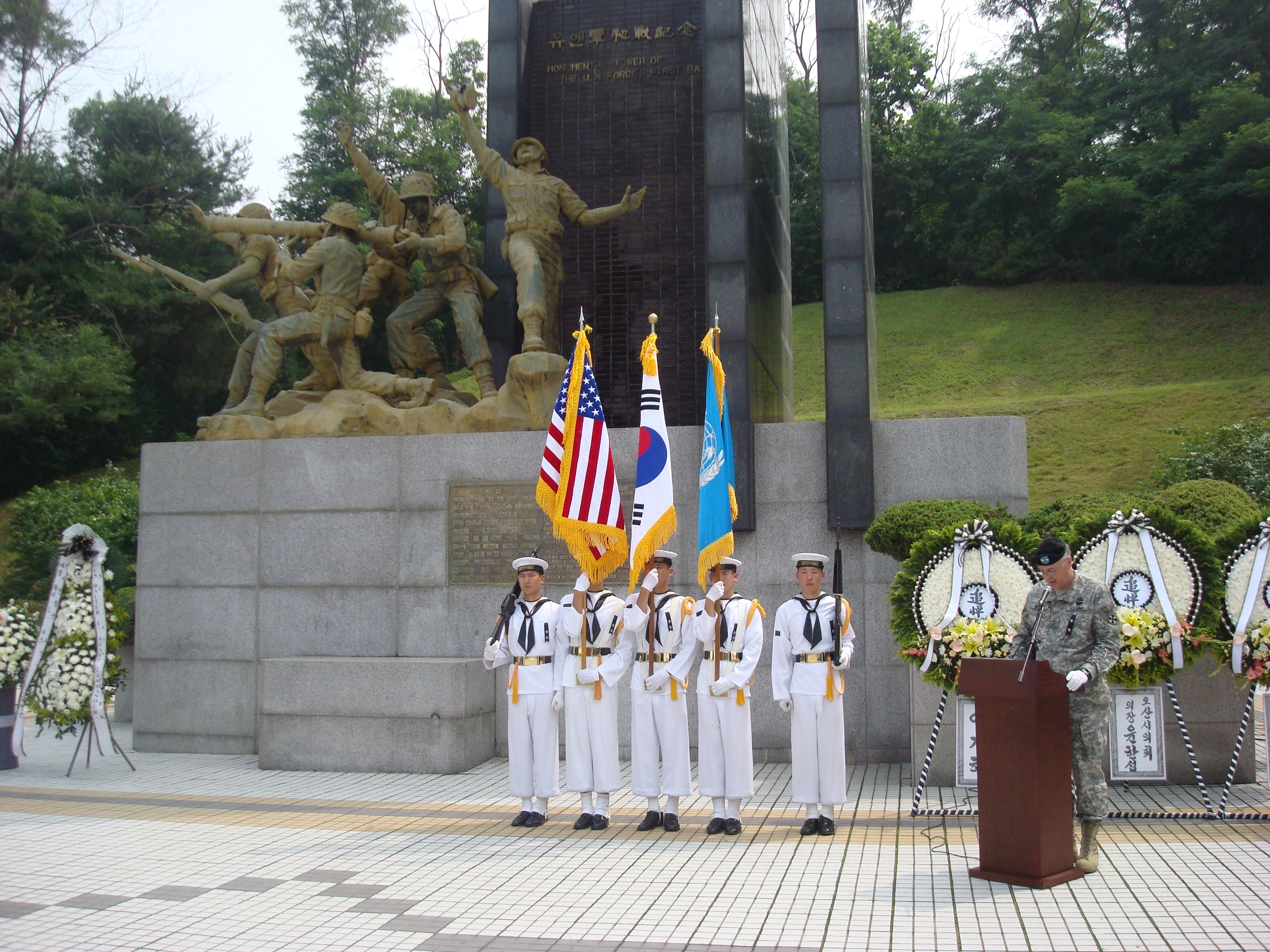 넓은 기념비 앞에 흰색 해군 제복을 입은 수병들이 줄지어 서 있고, 녹색 군복을 입은 다른 군인이 옆 연단에 서 있는 모습