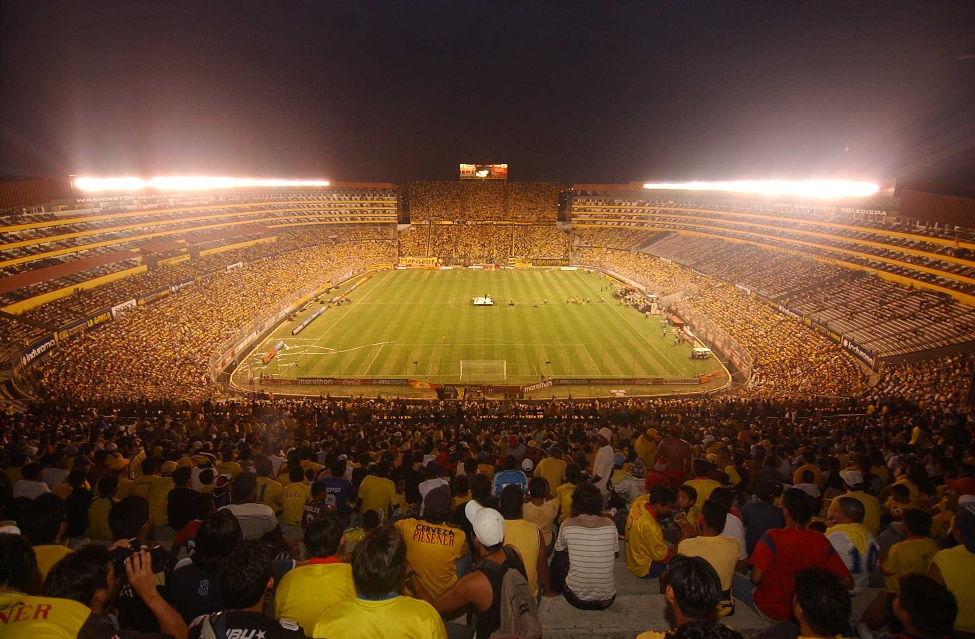 alt=Large football stadium with yellow seats and a green pitch.