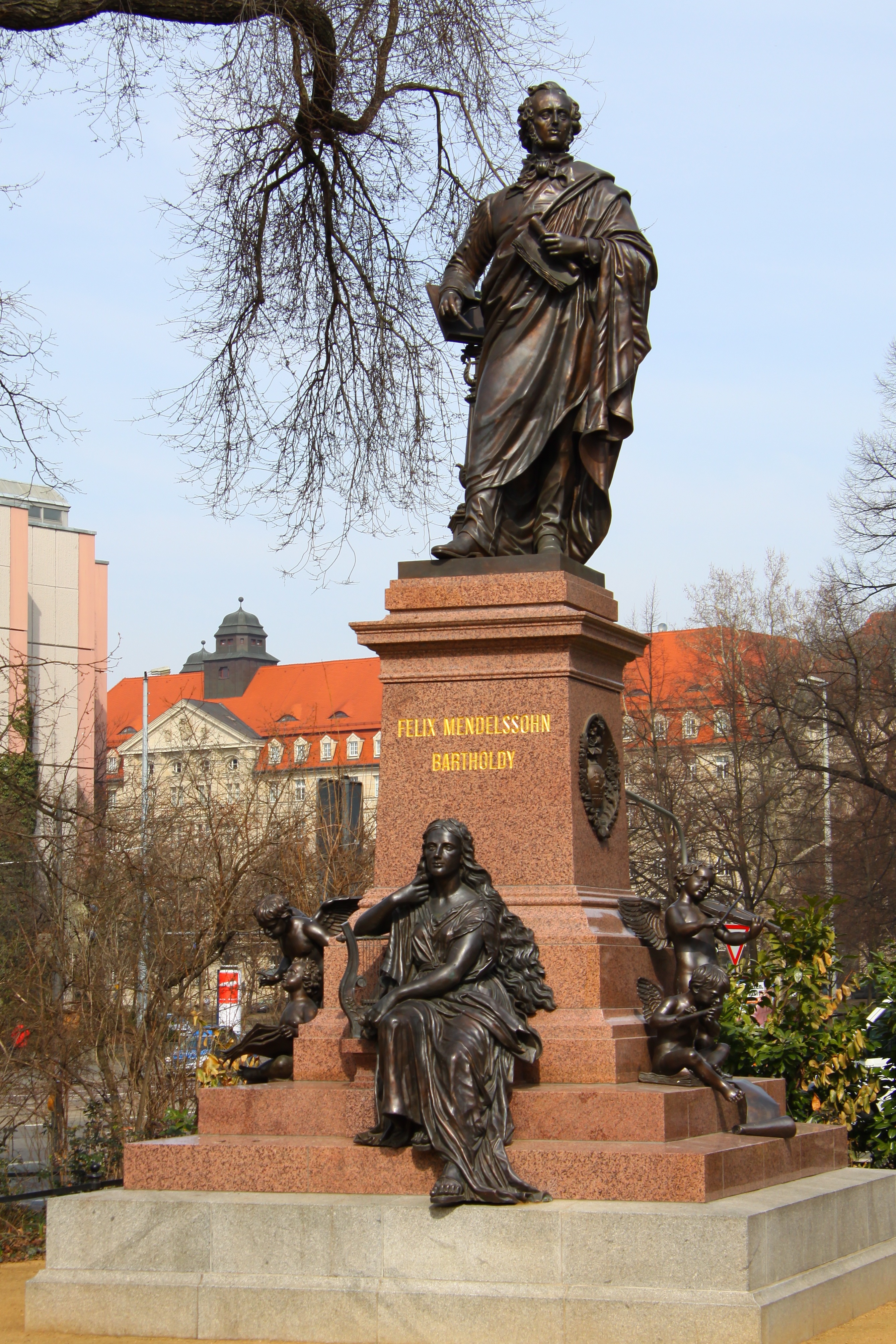 로브를 입은 남성상이 계단식 받침대 위에 있으며, 'Felix Mendelssohn Bartholdy'라고 새겨져 있다. 기둥 아래에는 현악기를 든 앉은 여성상이 있다. 야외 공간에 위치해 있다.