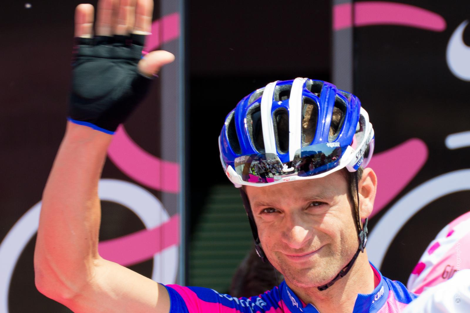 A cyclist being introduced to a crowd, waving his right hand in recognition