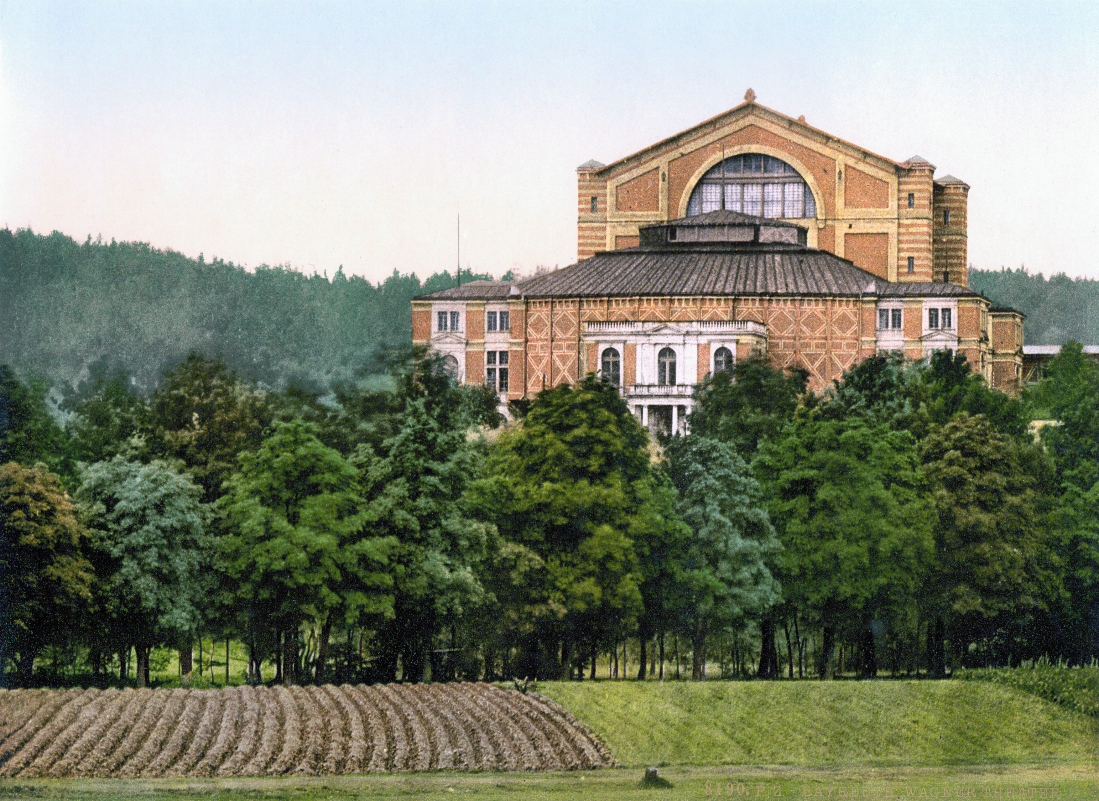A building stands beyond a part-ploughed field and a row of trees. It has five sections. Farthest away, the tallest part with a v-shaped roof contains the stage. Adjoining it is the auditorium section built of patterned brick. Nearest is the royal entrance, made of stone and brick with arched windows and a portico.