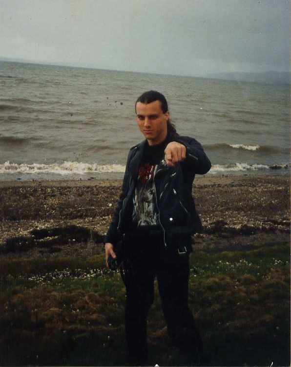 어두운 해변가에 서 있는 척 슐디너(Chuck Schuldiner)의 사진. 그는 긴 머리에 검은색 바지, 검은색 셔츠, 그리고 검은색 가죽 재킷을 입고 있습니다.