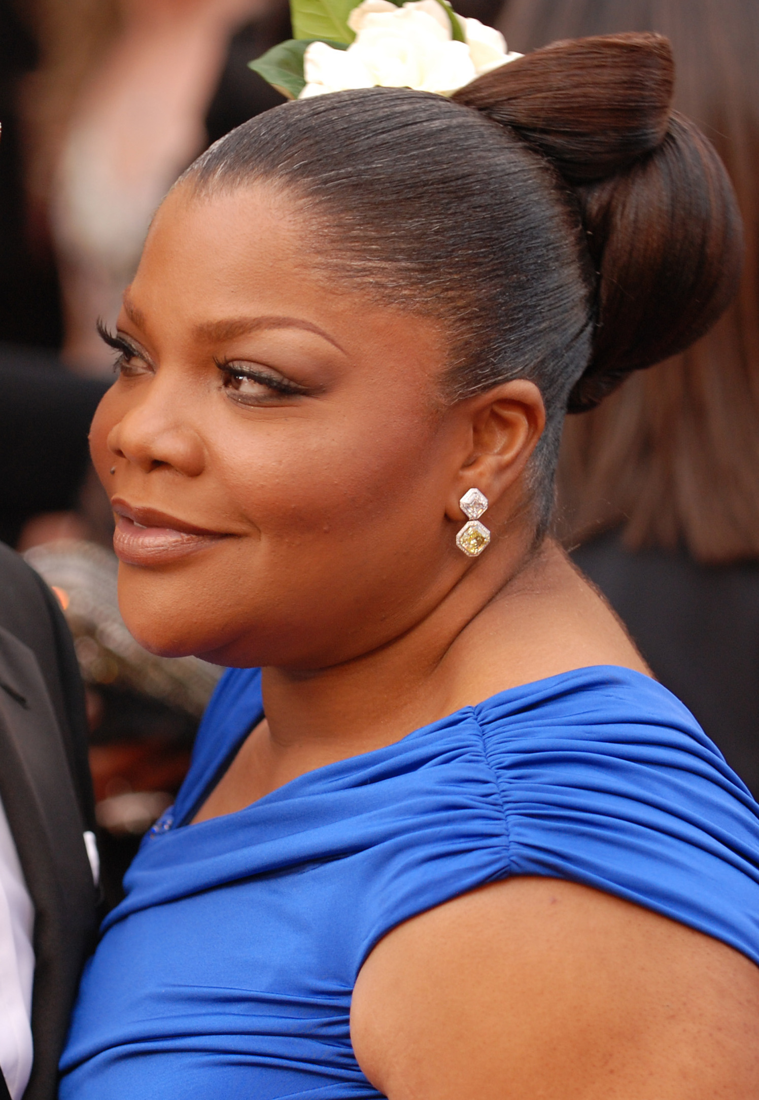 An African American female with dark brown hair up in a bun with a flower. She is wearing a short sleeved royal blue dress.