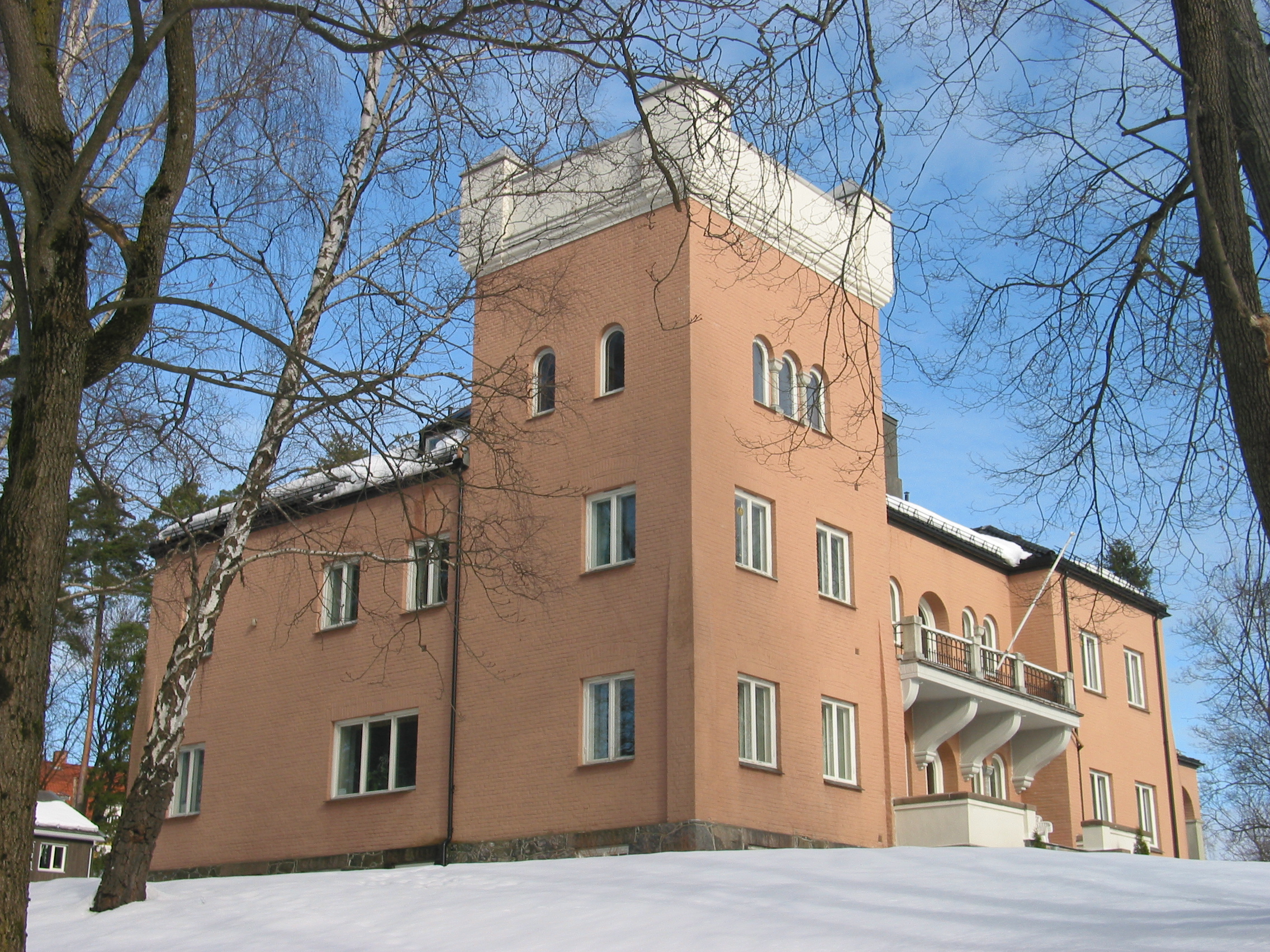 A substantial building in pink stone or cladding, consisting of a three-storey square tower with two two-storey wings. The building stands, framed by trees, in a snow-covered garden.