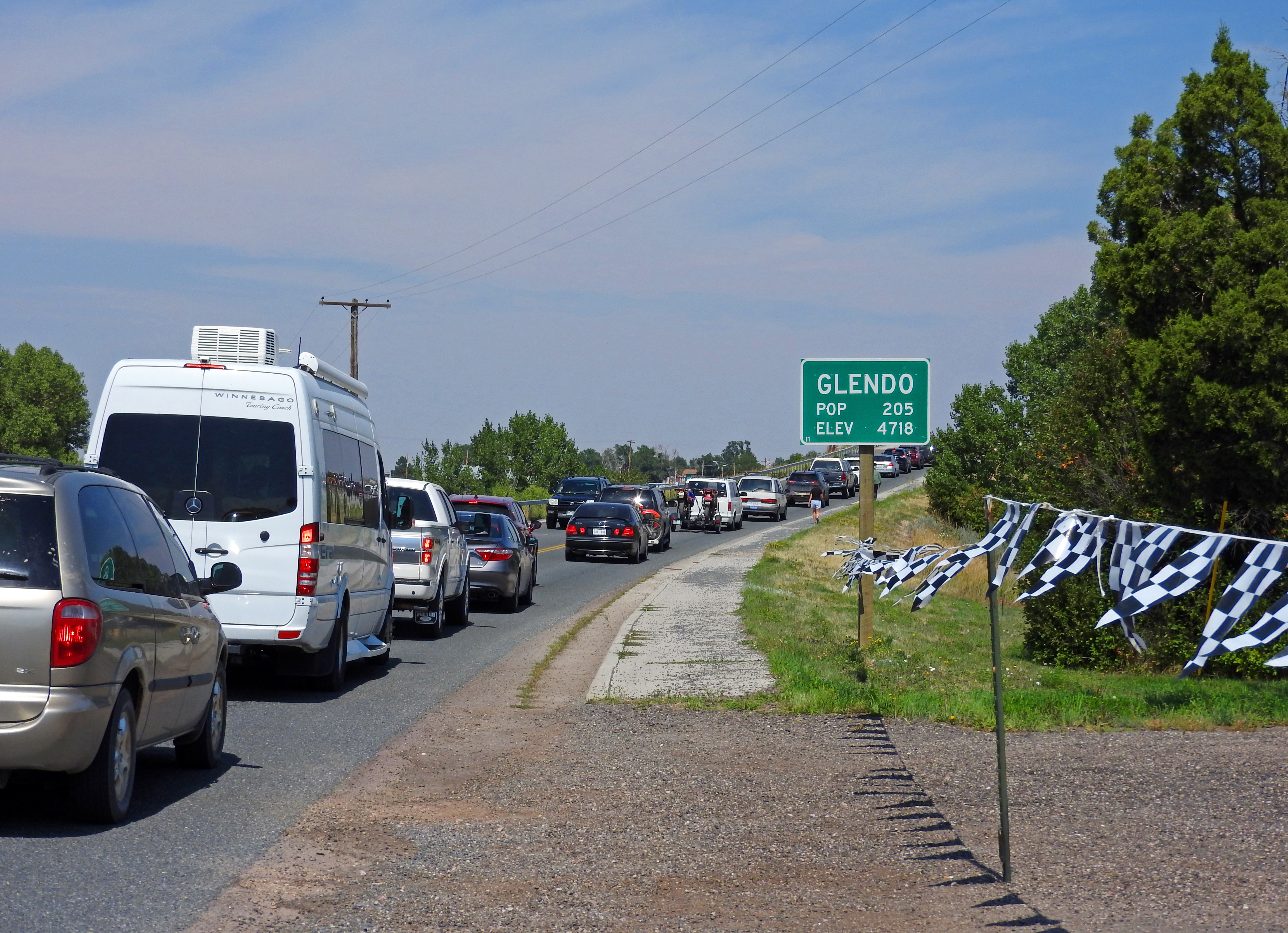 A roadway curving slightly to the right around some tall trees with cars stopped on it. To its right midway through the image a white on green sign says