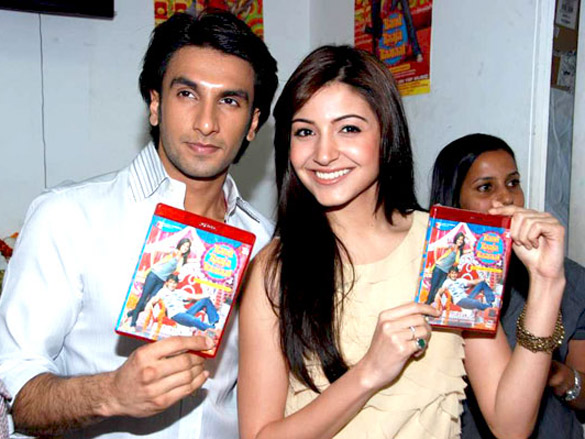 Anushka Sharma and Ranveer Singh holding up DVDs. While Sharma, wearing a pale yellow top is smiling directly at the camera, Singh, dressed in a white shirt, is looking away.