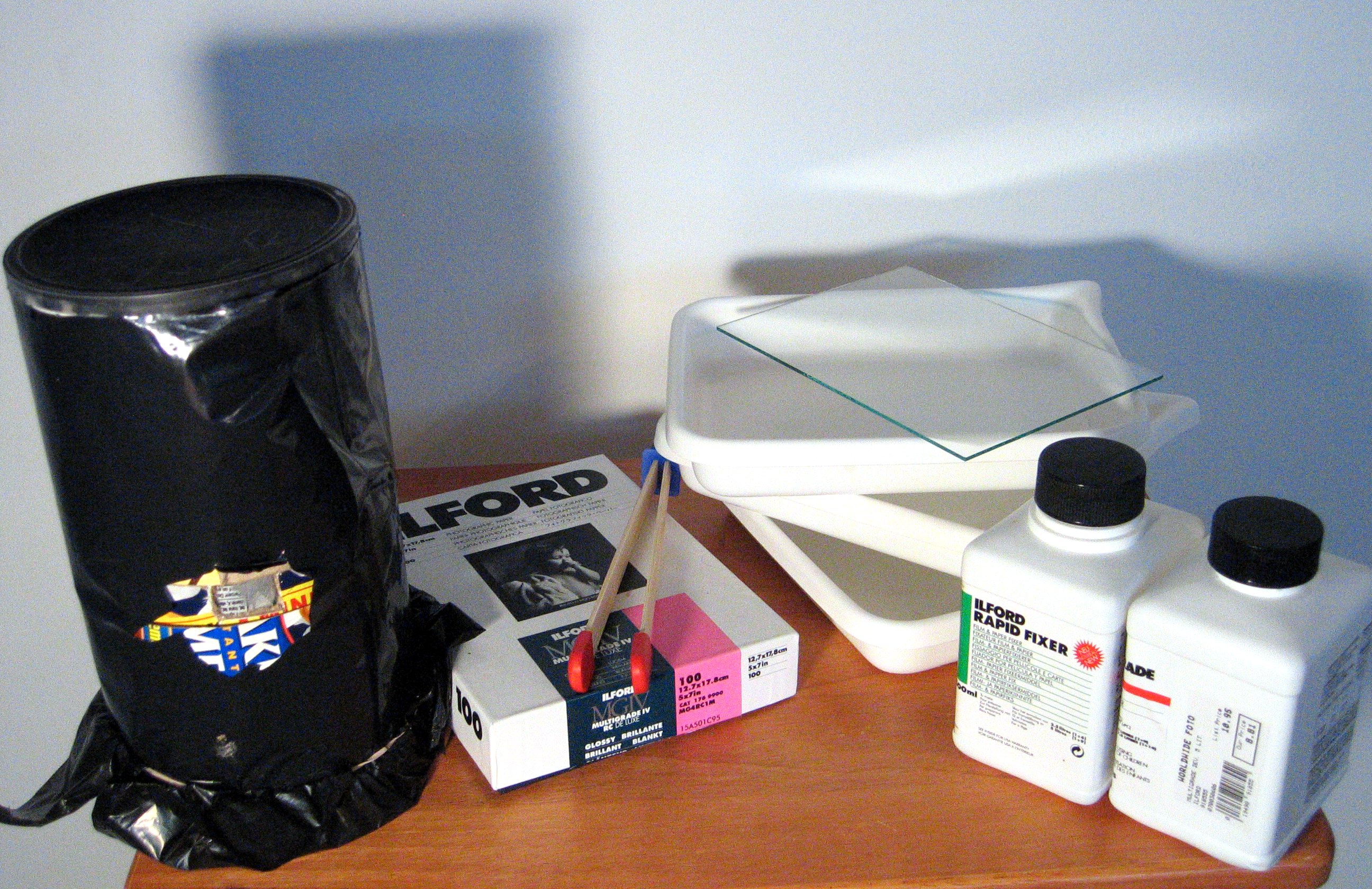 A pinhole camera made from an oatmeal container, wrapped in opaque plastic to prevent light leaks; a box of photographic paper; tongs and dishes for developing film; bottles of film developing chemicals