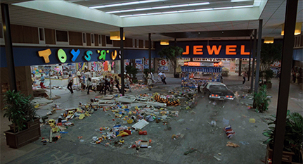 Film screenshot showing a police car driving through a shopping mall: Scattered items are present on the floor and people are running away from the vehicle. Stores visible in the mall include Toys 