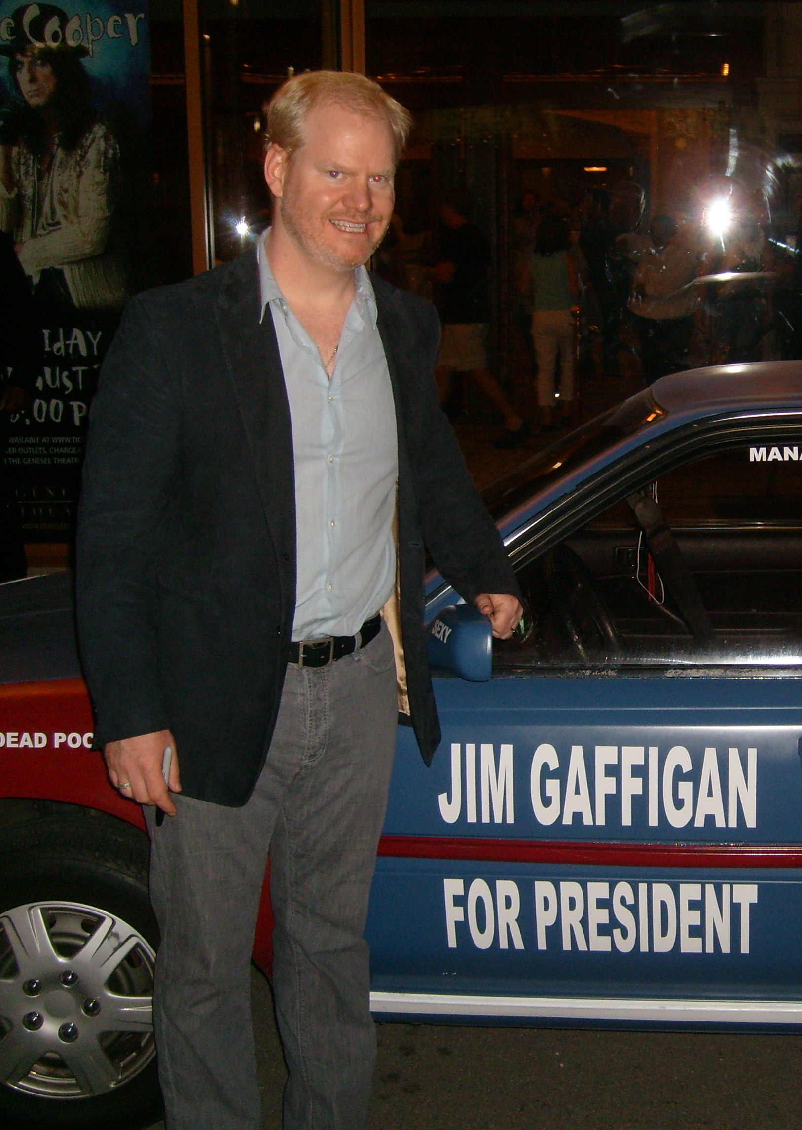 Gaffigan standing next to a car reading 