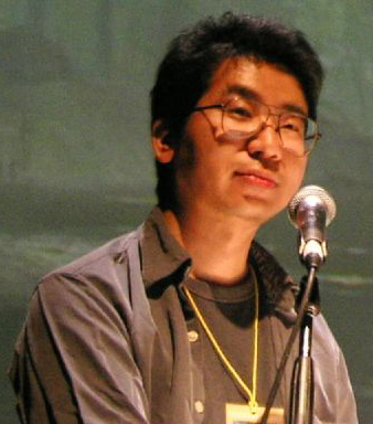 검은 티셔츠와 회색 재킷을 입은 안경 쓴 일본 남자가 기대에 차서 앞을 바라보고 있다.