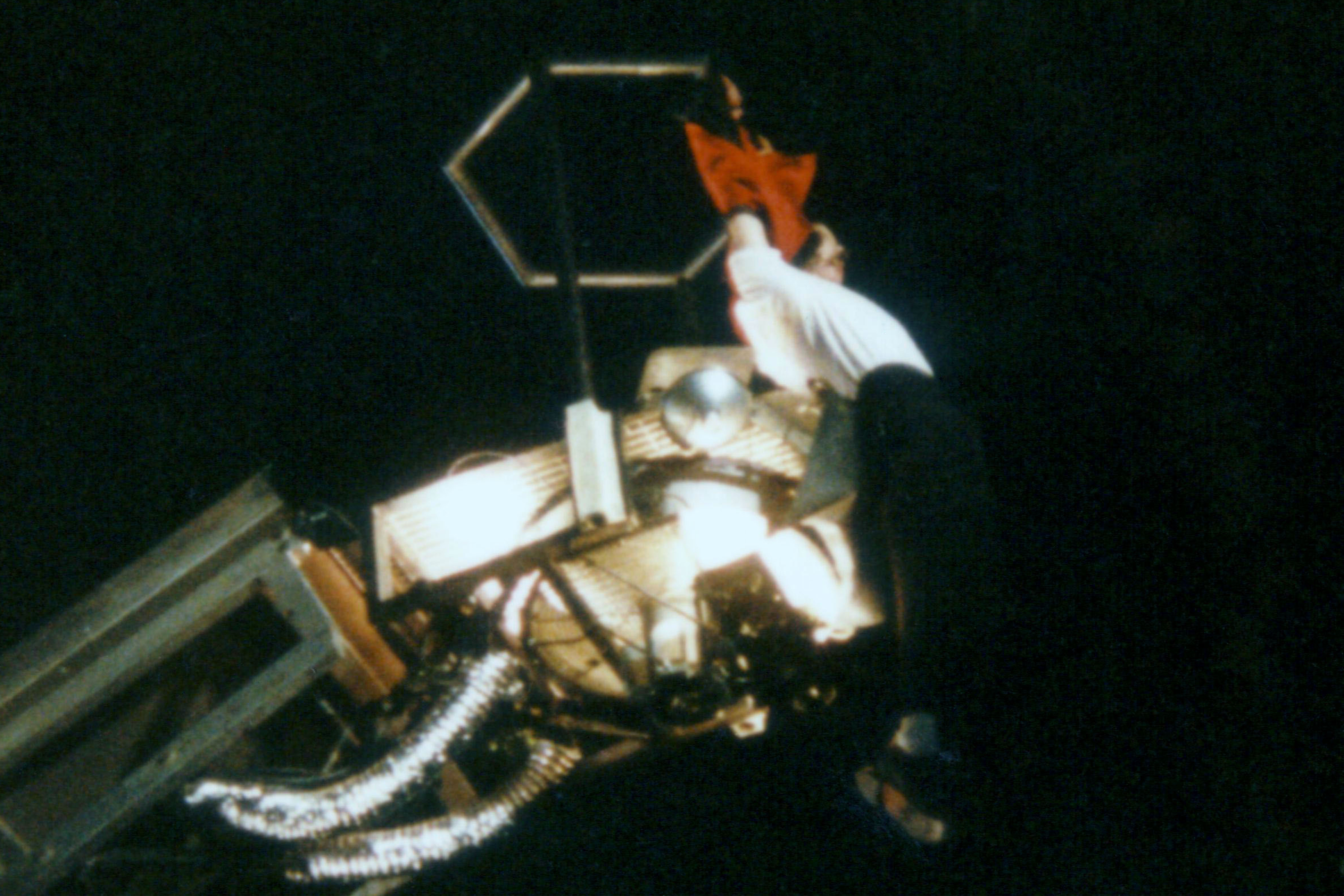 A image of a person with pale skin dangling by their hands from a crane. The person is wearing a white shirt and black pants and footwear. A black background can be seen behind the person.
