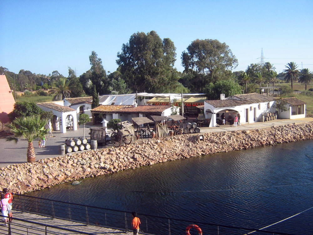 Colour photo showing the reconstruction of the ancient port of ''Palos de la frontera'': transportation barrels, low buildings bordered by a covered gallery, a chariot at the very edge of the water awaiting the arrival of a boat.