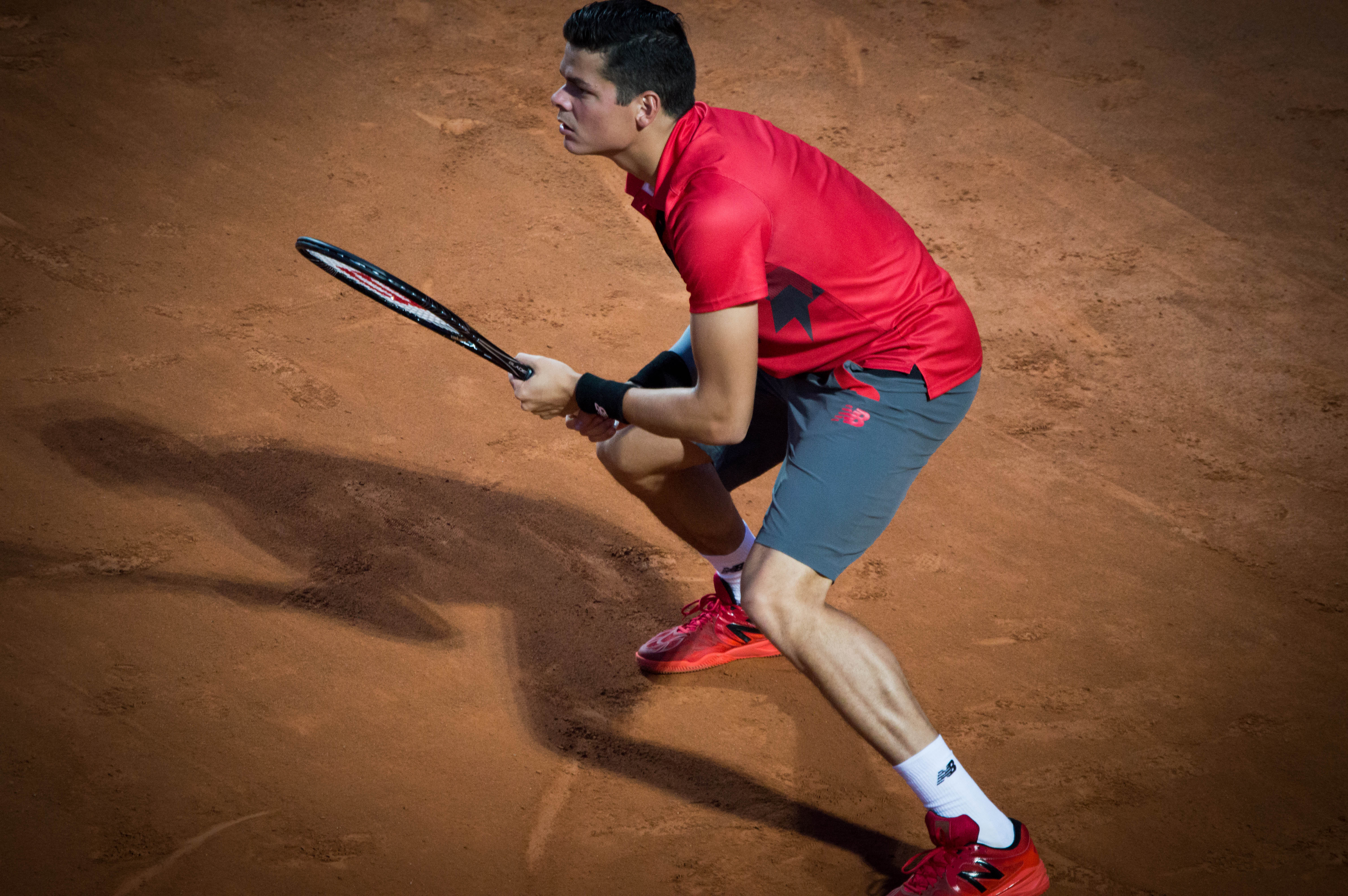 Raonic standing on clay, with his feet wide apart, both hands on his racquet, and looking left. His shadow stretches out sharply to the left.