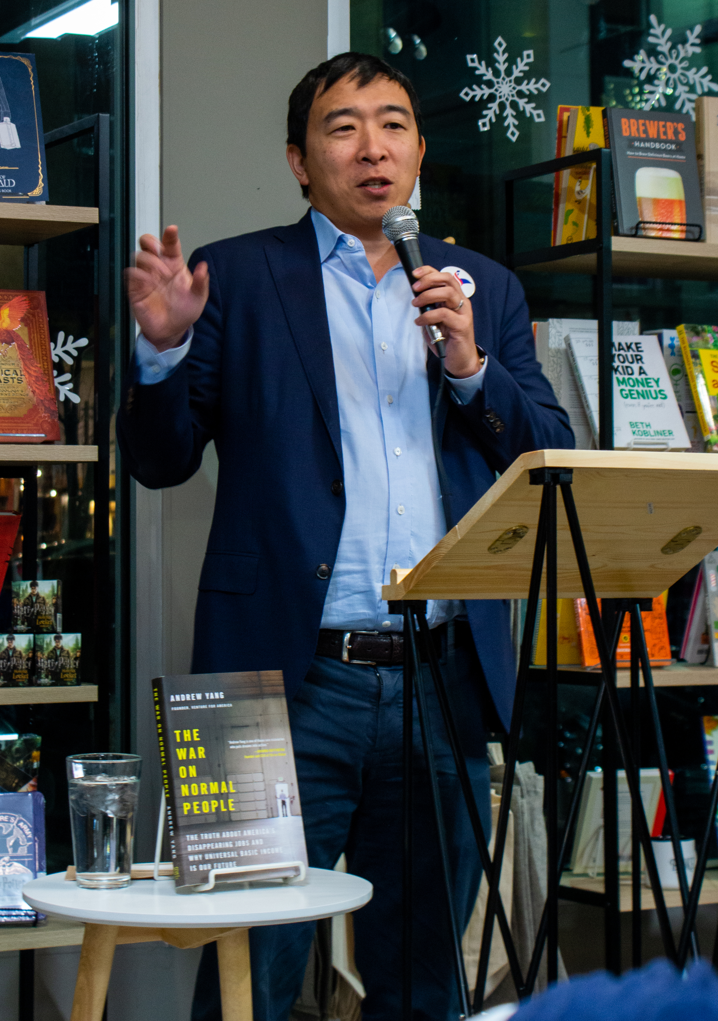 Yang is holding a microphone while gesturing and making a speech. His book, ''The War on Normal People'', is displayed on a table in front of him.