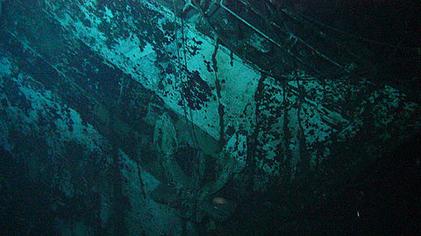 Close up photograph of part of a ship's bow. The bow is at an angle. An anchor is located in the centre of the image. A coloured band runs from the top left corner to the right third of the bottom edge. A dark square and the remains of a number