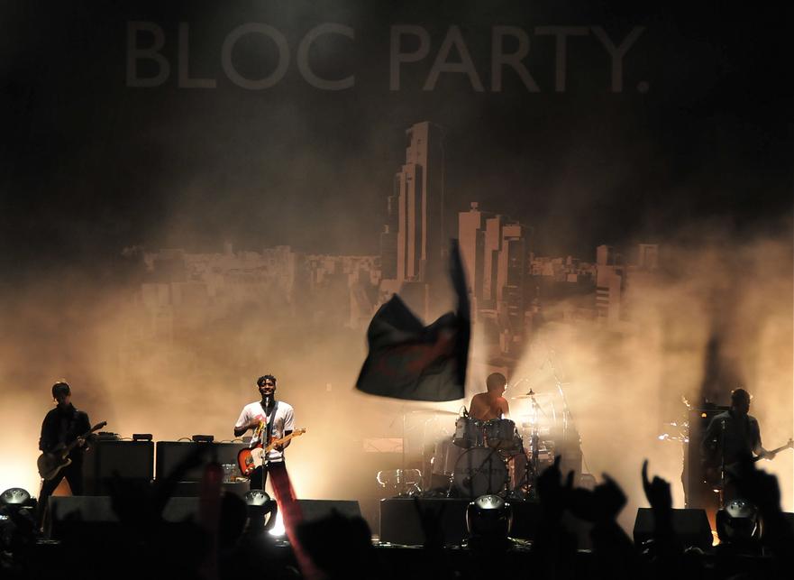 Two guitarists, a drummer, and a bassist are performing on a smoky stage lit by white stage lights. The stage background is a black tarpaulin emblazoned with 