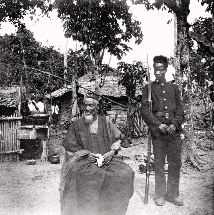Pemimpin Temne Bai Bureh terlihat di sini pada tahun 1898 setelah penyerahannya, duduk santai dengan pakaian tradisionalnya dengan saputangan di tangannya, sementara seorang prajurit Pasukan Perbatasan Afrika Barat Sierra Leone berjaga di sebelahnya