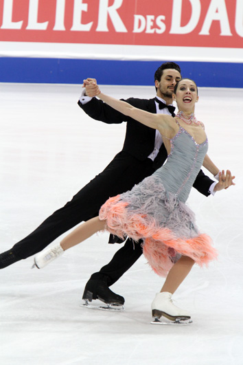 A man and woman ice dancing; the man, on the left, is wearing a classic tuxedo with a white-ruffled shirt, and the woman is wearing a knee-length light blue dress with a ruffled pink hem 